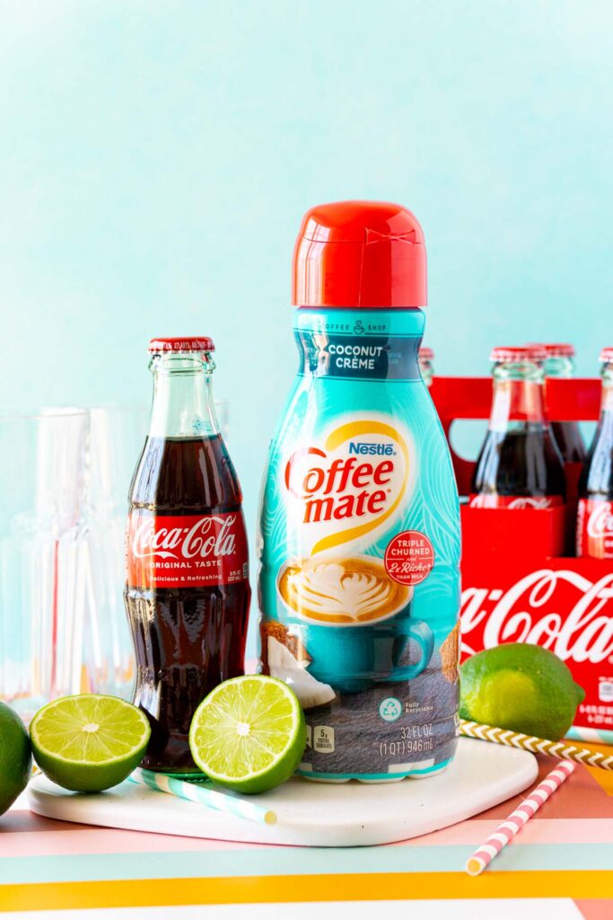 A bottle of Coca-Cola next to Coffee Mate coconut creamer and fresh limes, representing the core ingredients of a ready to drink dirty soda concept.